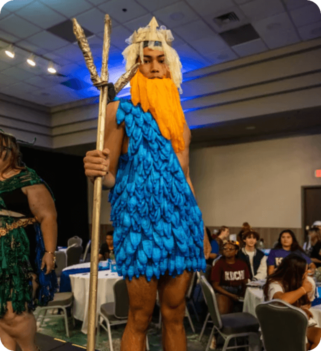 Person in a blue costume with a beard and staff at an event with tables and people in the background.