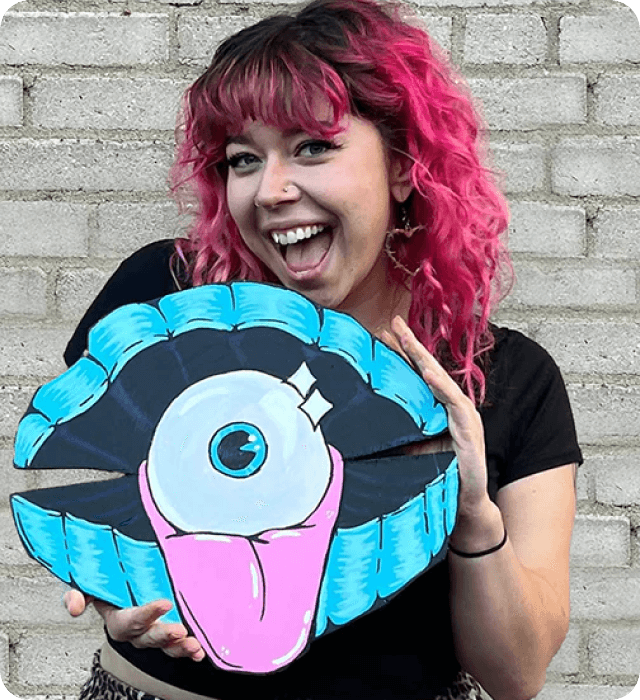 Person with pink hair holding a colorful, eye-shaped object against a brick wall.