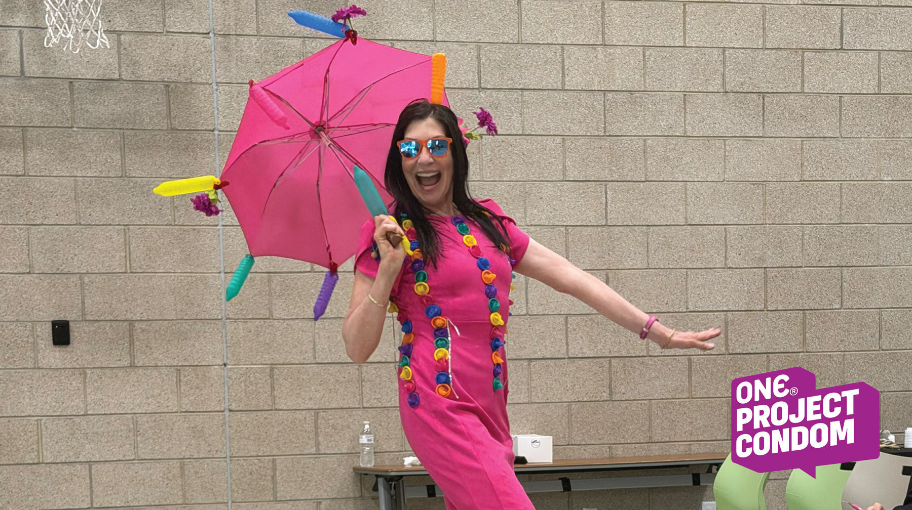 Fashion show model posing on the runway in a bright pink dress lined with colorful condoms and carrying a matching statement umbrella.